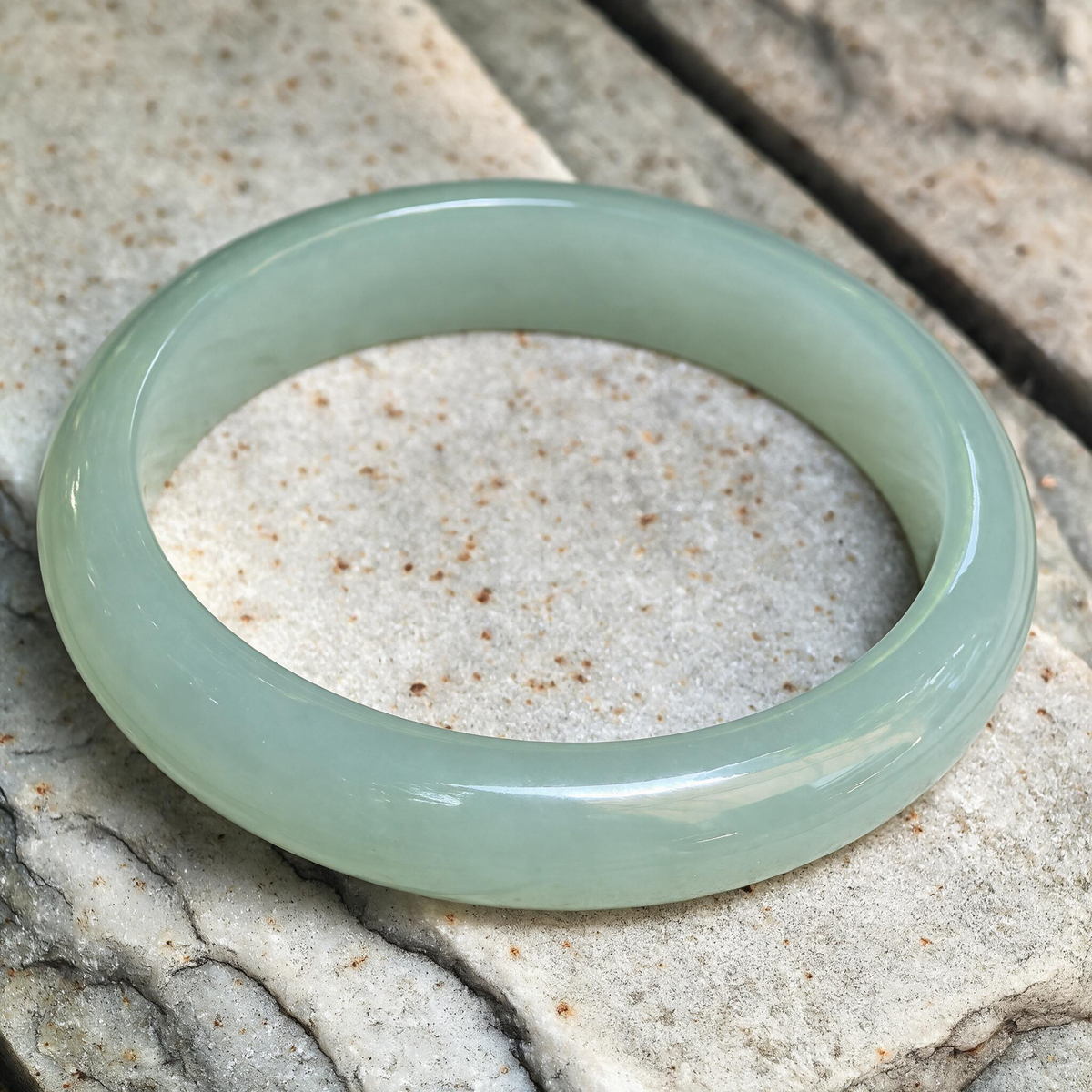 Angled view of a light green icy glutinous jadeite bangle resting on stone, showing its flatter inner face and rounded outer profile.