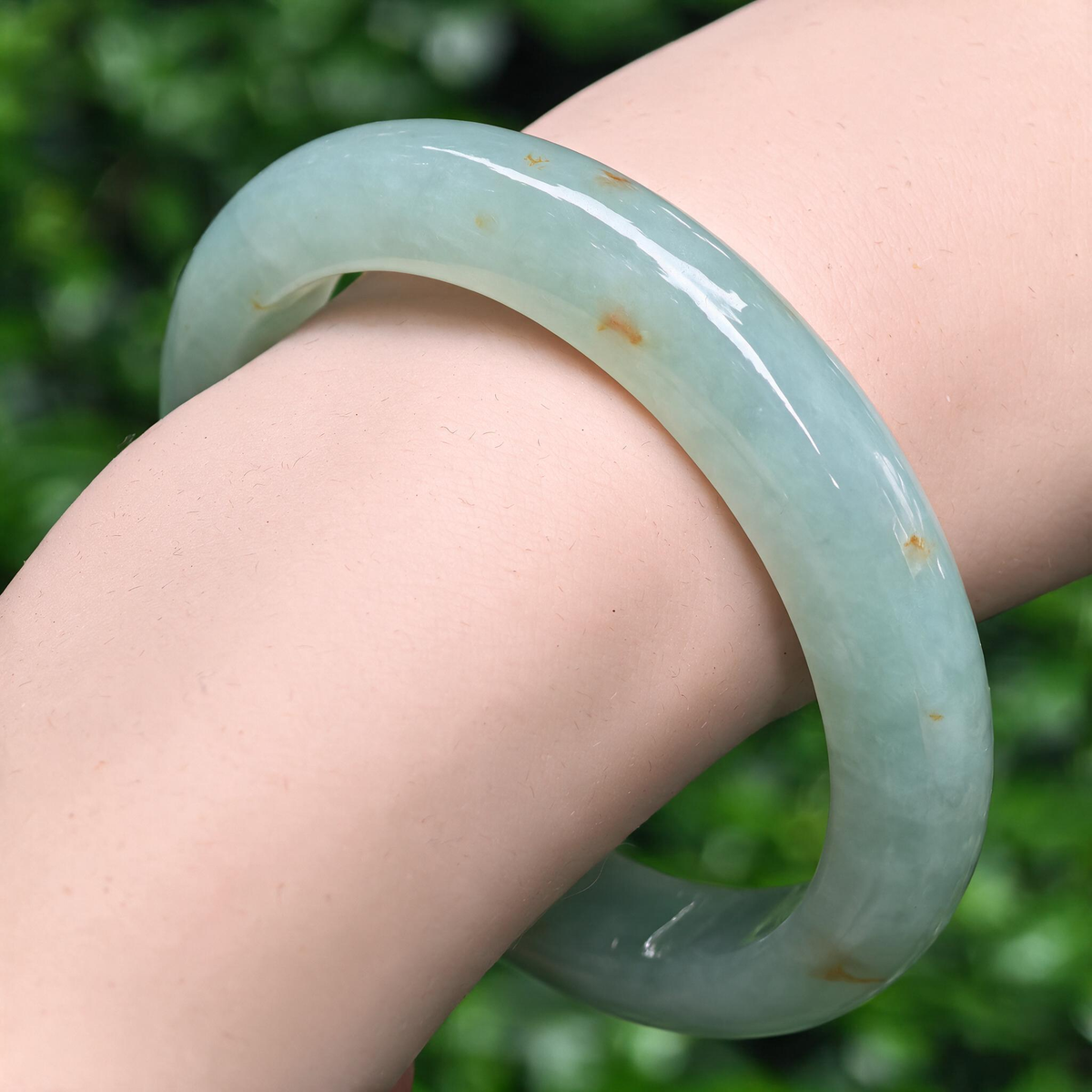 Angled wrist view of the pale green jadeite bangle, showing its rounded outer profile and scattered natural golden-brown flecks.