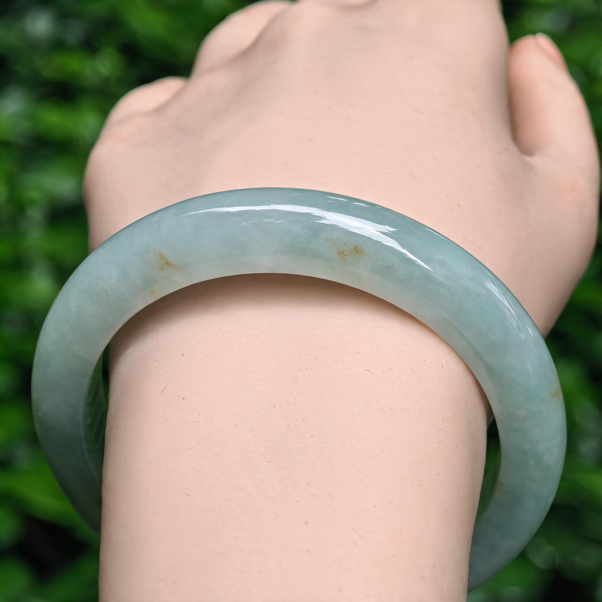 Front wrist view of a pale apple green jadeite D-shape bangle with small golden-brown flecks against an outdoor leafy background.